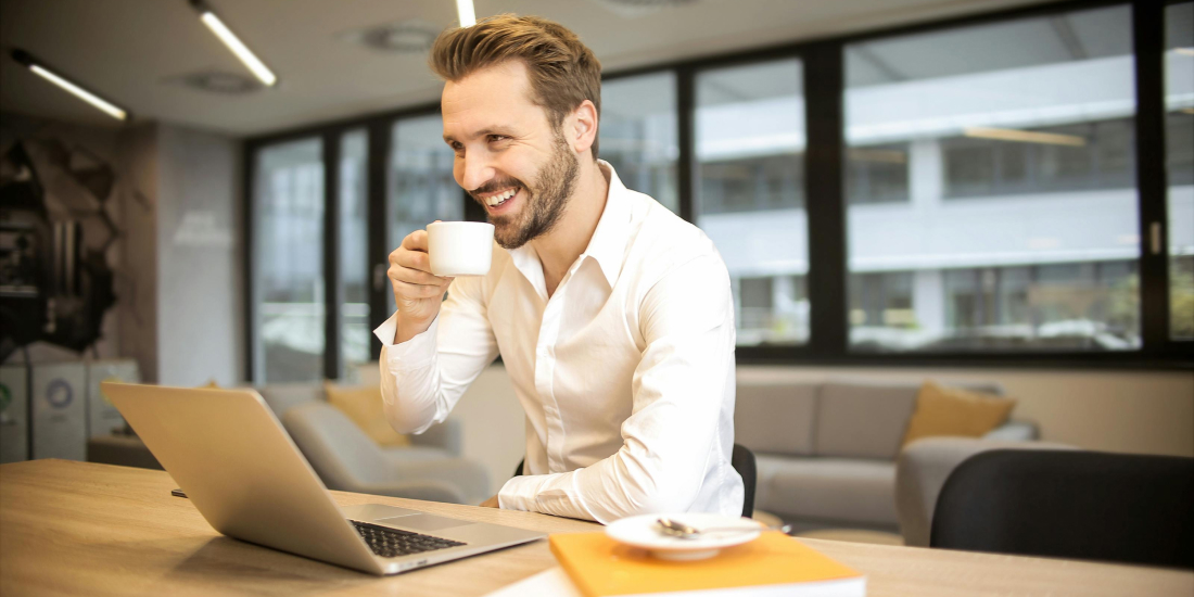 Estate agent drinking a tea and smiling. A laptop is on the table in front of him.